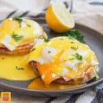 Eggs benedict on a plate: two poached eggs over slices of ham on English muffins, on a black plate with half a lemon and fresh herbs