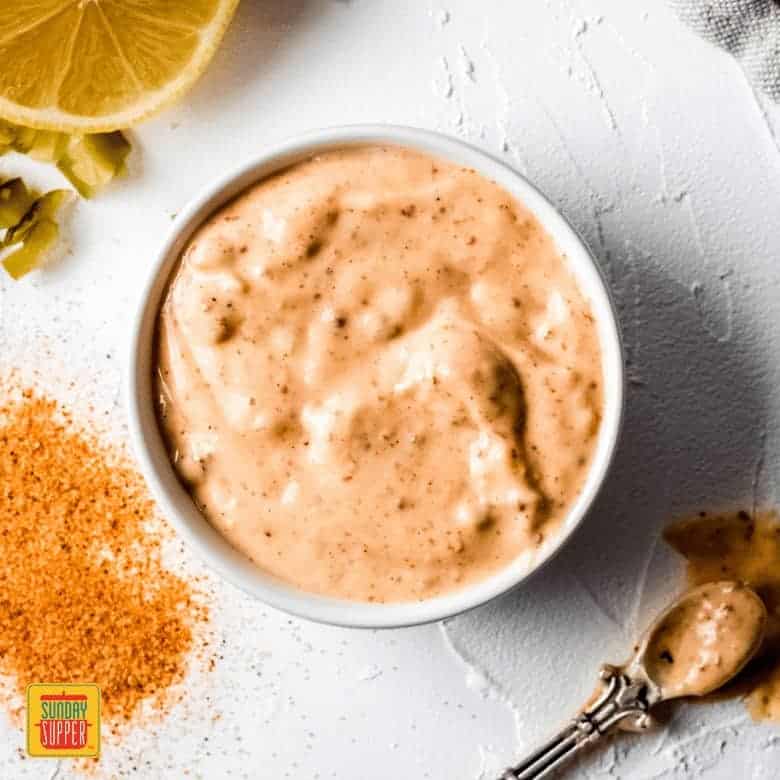 a small white bowl of remoulade sauce. There are ingredients laying on the table around the bowl and a small stirring teaspoon laying next to the bowl.