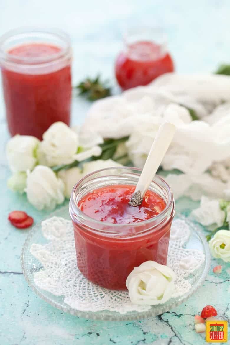 Strawberry rhubarb jam in a small jar with a serving spoon, surrounded by white roses