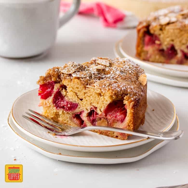 A slice of rhubarb cake on a plate with a fork next to it and mug in the background