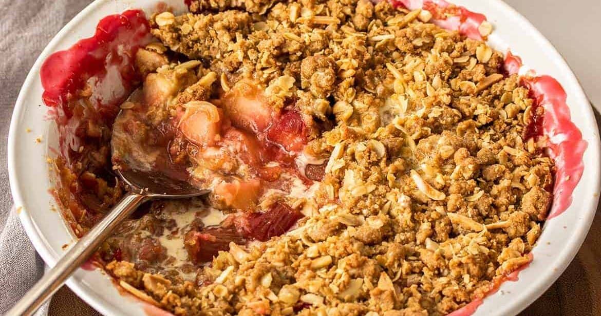 A white pie dish filled with a fruit crumble with a spoon in the side