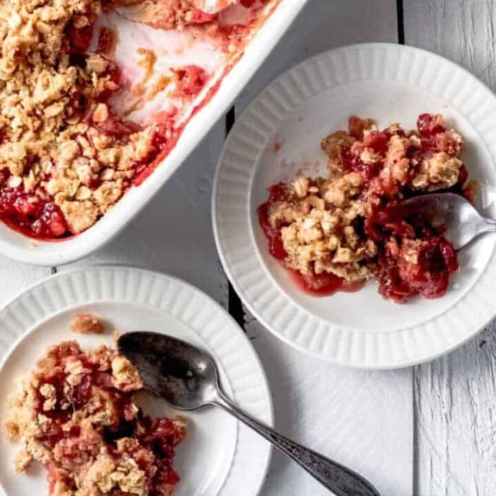 two servings of strawberry rhubarb crisp on small white plates. There is a 9x9 dish sitting beside the plates.
