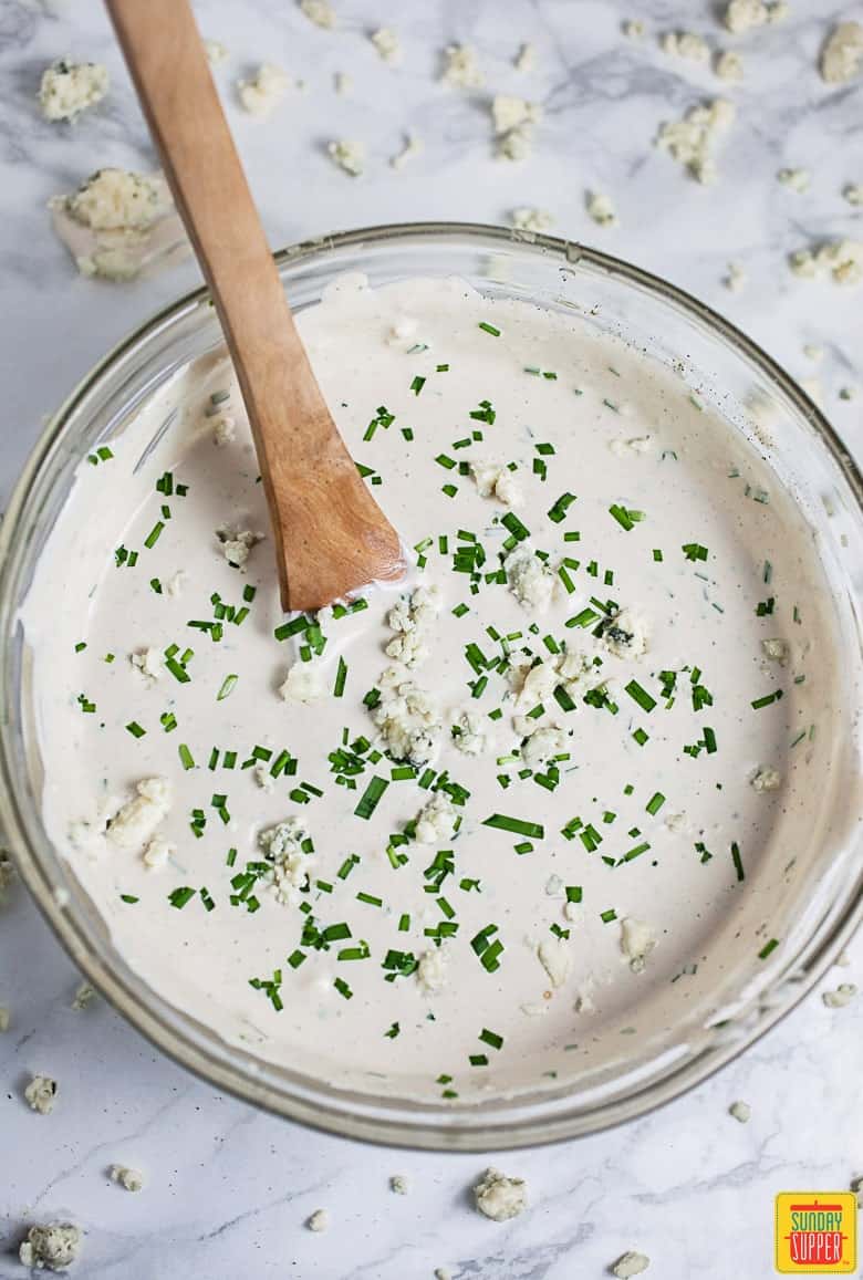 How To Make Blue Cheese Dressing: dressing in a bowl with a wooden spoon and blue cheese crumbles