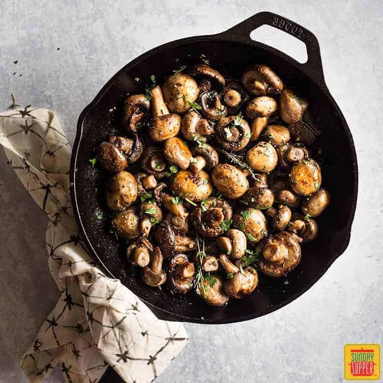 Button mushrooms in a cast iron skillet, cooked and ready to serve, the handle of the skillet is wrapped in a towel with a hexagon pattern