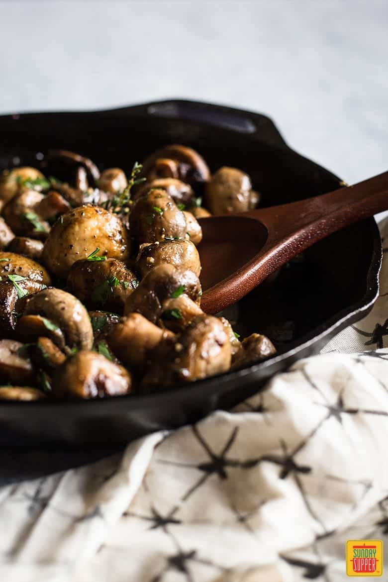 Button mushrooms in a cast iron skillet being scooped with a wooden spoon
