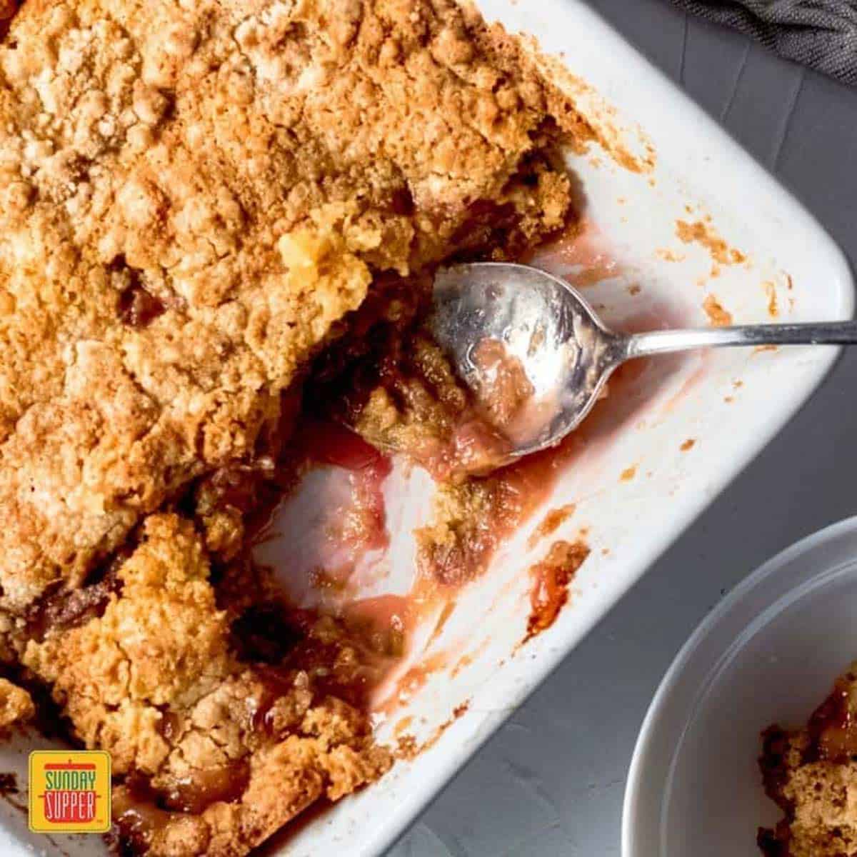 Rhubarb dump cake in a white dish with a serving spoon and a bowl of dump cake to the side