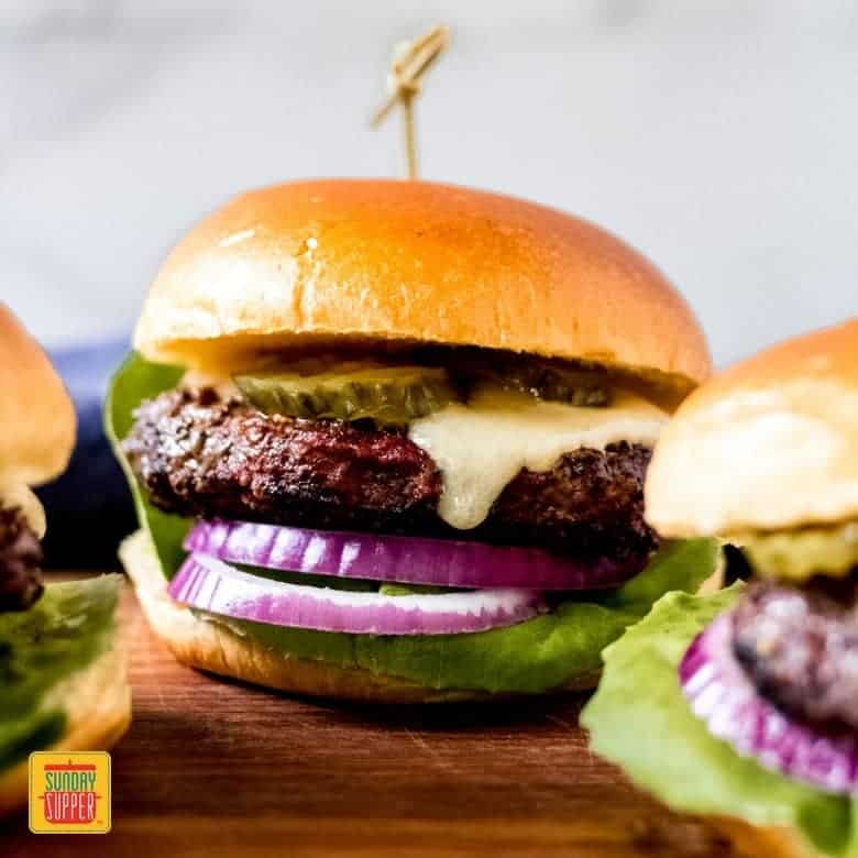 Close up of a bison burger surrounded by two other bison burgers on a bun with pickles, onions, lettuce, and cheese melting down the side