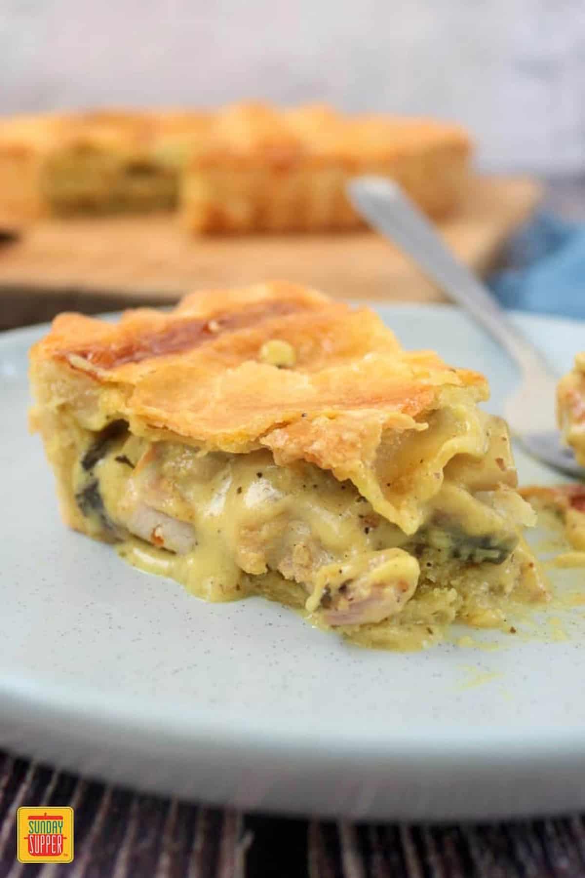 A slice of chicken pie on a light gray plate with a fork and the whole pie blurred in the background