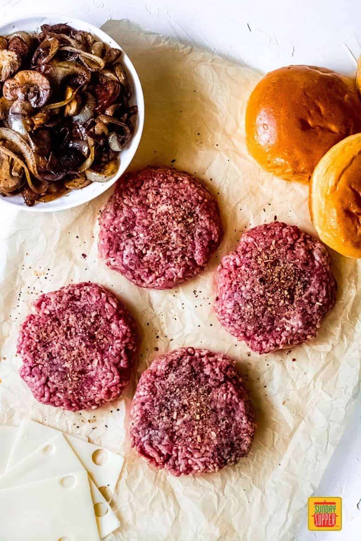 Four ground beef hamburger patties on parchment paper with four slices of swiss cheese, a bowl of sautéed mushrooms and onions, and brioche buns, ready to make Mushroom Swiss Burgers