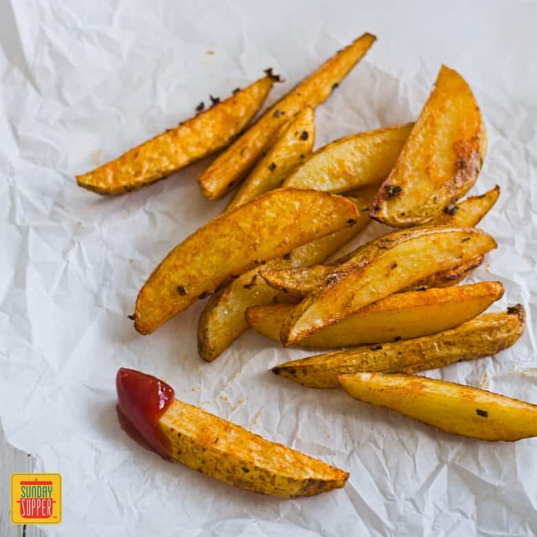 baked crispy potato wedges served with ketchup on parchment paper, one is dipped in ketchup