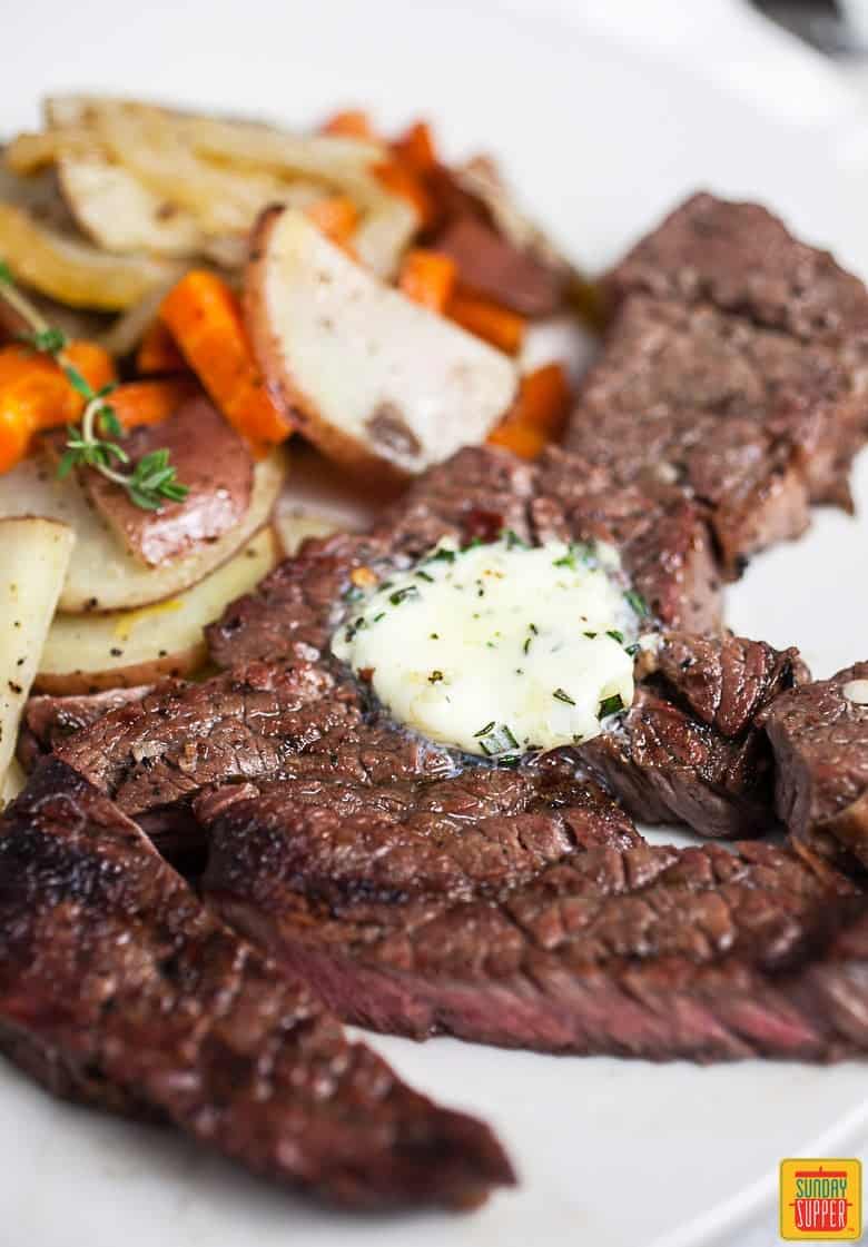 Close up of grilled chuck steak on a white plate with a side of potatoes and carrots and topped with garlic butter
