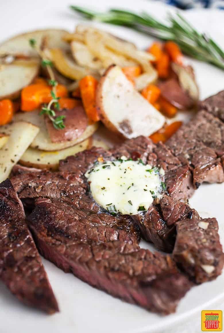 Sliced grilled chuck steak with garlic compound butter on top on a white plate next to vegetables