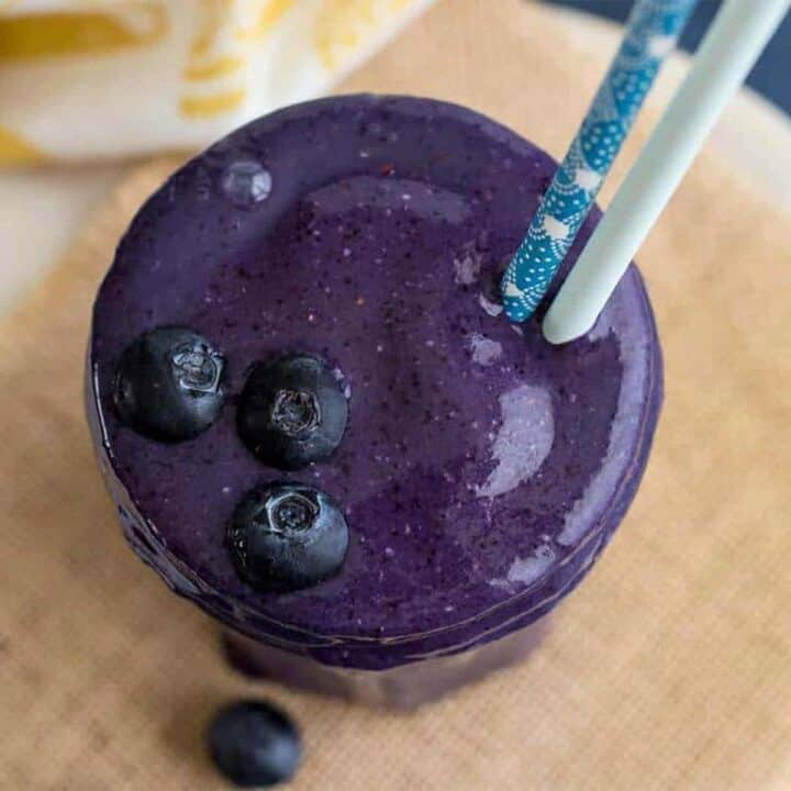 Top-down photo of a blueberry smoothie in a glass with two straws and fresh blueberries on top