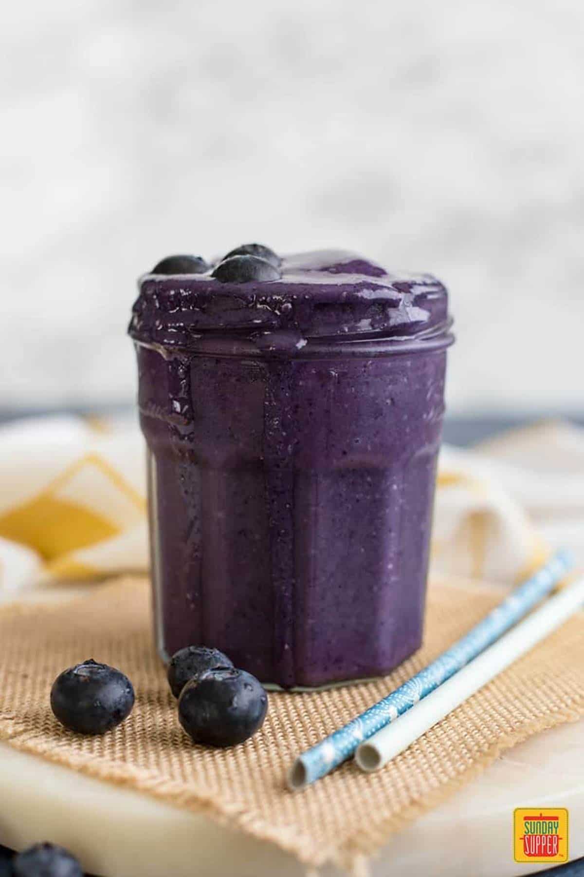 Blueberry smoothie in a glass next to two straws and three fresh blueberries