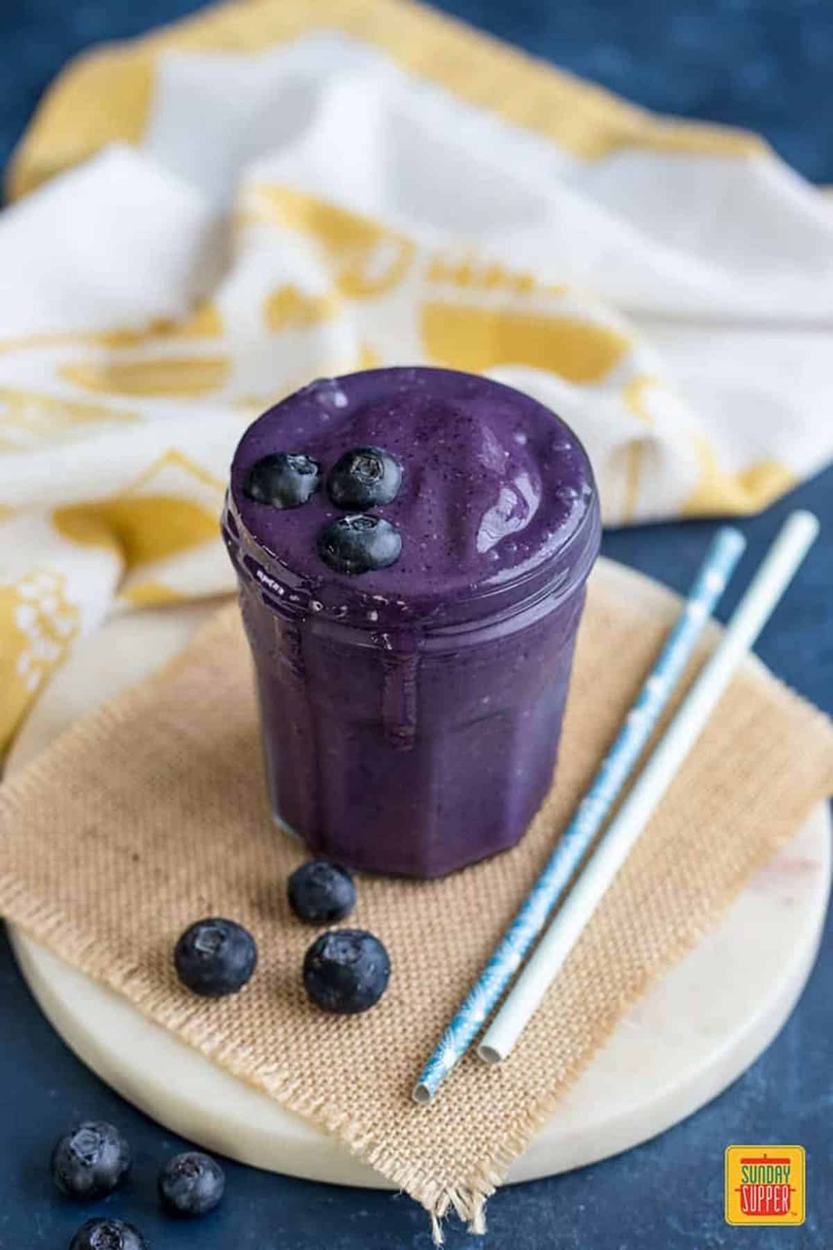 A glass jar filled with blueberry smoothie next to three fresh blueberries and two straws