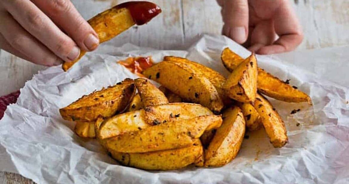 A plate of baked crispy potato wedges, holding one dipped in ketchup