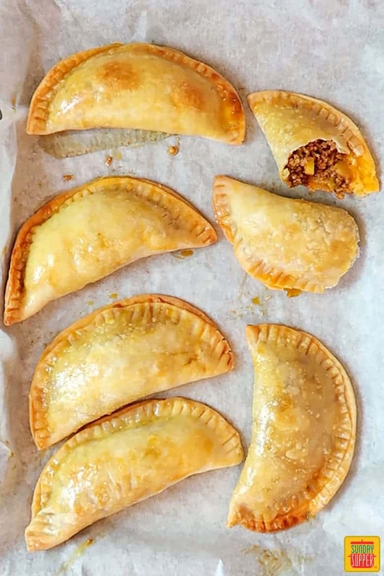 Baked empanadas on a baking sheet with one split open and ready to eat