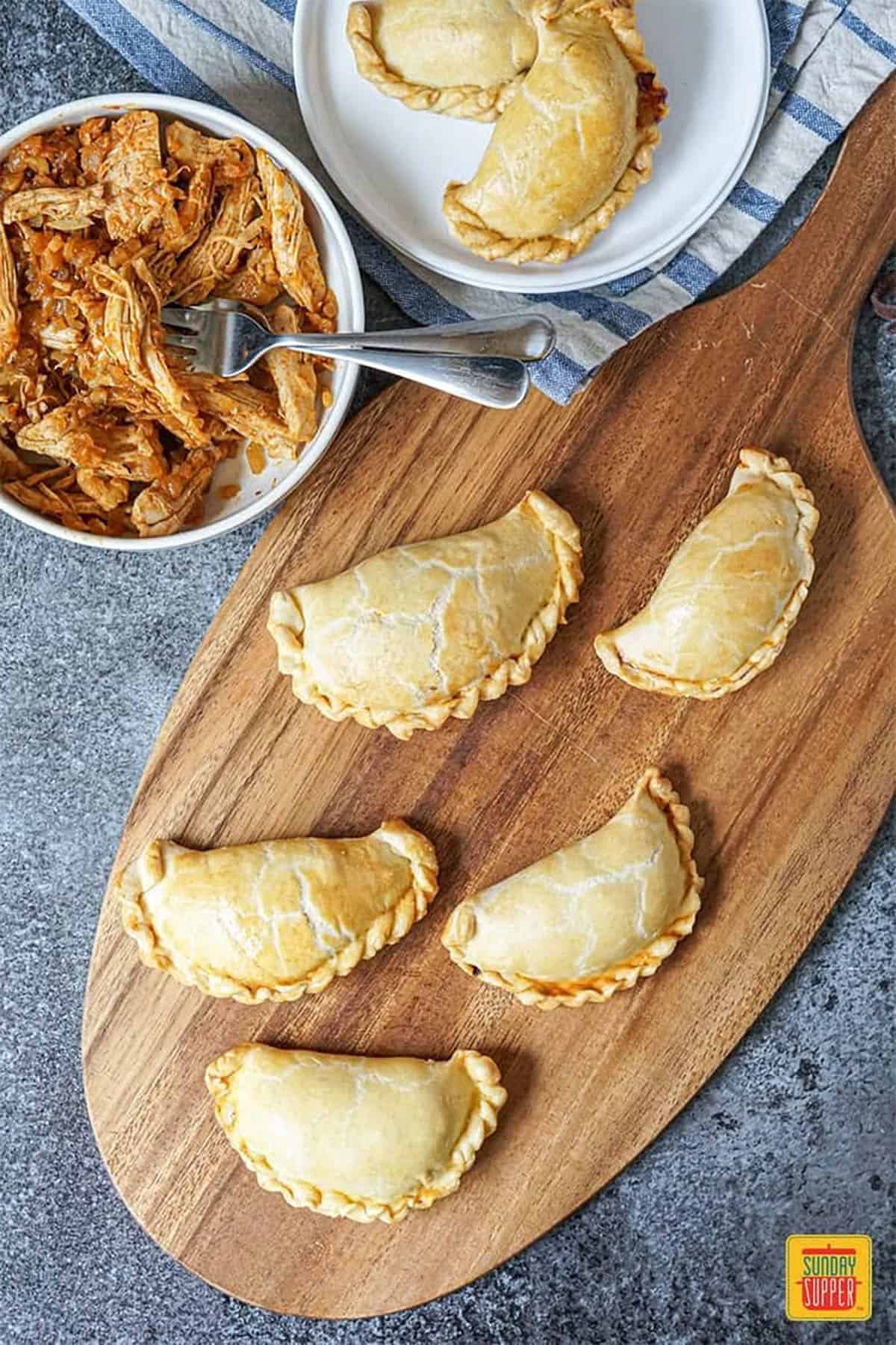 Baked chicken empanadas on a wooden serving board next to a bowl of shredded chicken filling