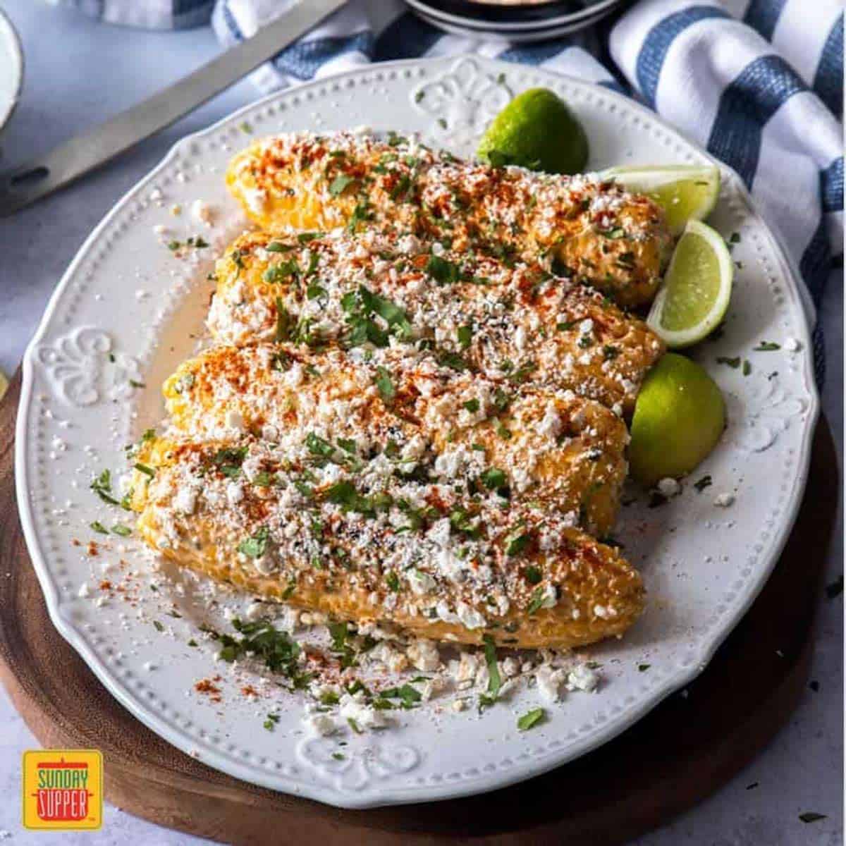 Four grilled corn on the cob on a white platter with Elote recipe topping and lime wedges