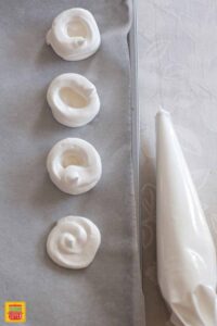 Pavlova shells piped onto parchment paper next to a piping bag full of pavlova meringue