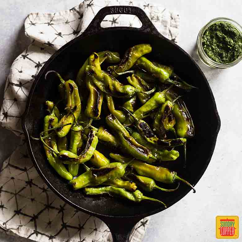 Blistered shishito peppers in a black cast iron skillet with a kitchen towel and citrus pesto off to the side