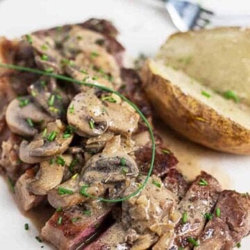 Steak diane recipe sliced on a white plate next to a baked potato cut in half