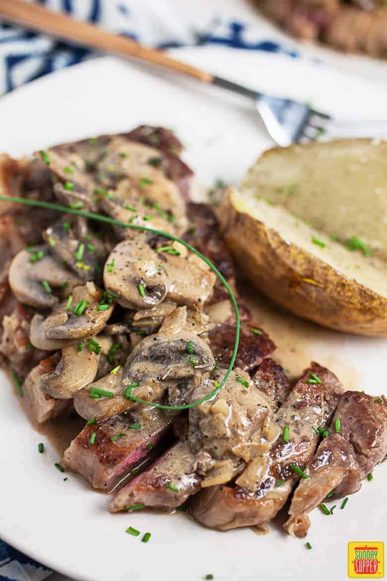 Steak diane recipe sliced on a white plate next to a baked potato cut in half