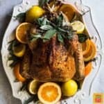 A whole garlic butter turkey on a white platter surrounded by fresh citrus and herbs