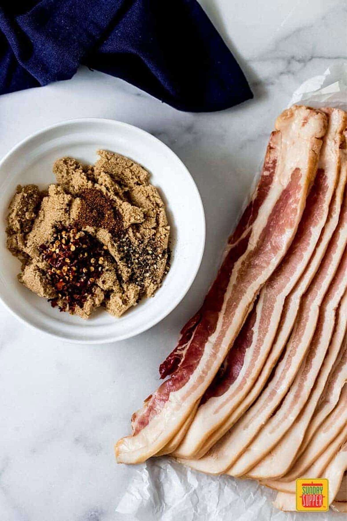 ingredients for candied bacon on a table - thick-cut bacon, and a bowl of brown sugar, cayenne pepper, black pepper, and red pepper flakes mixed together