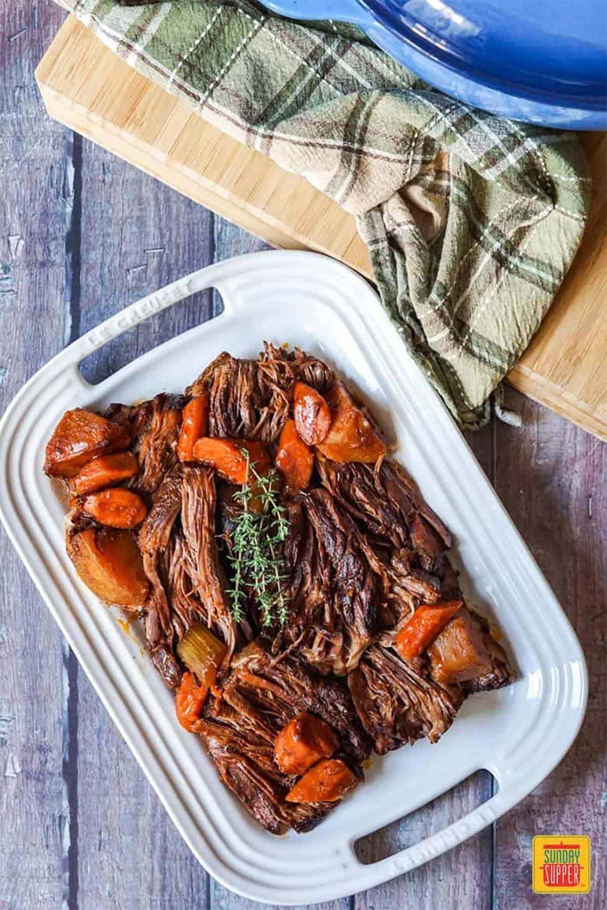 Shredded beef chuck roast with vegetables on a white platter next to the dutch oven lid