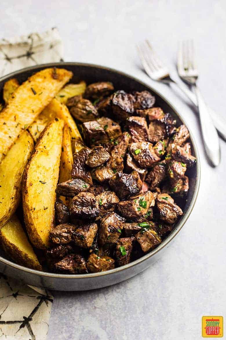Garlic Butter Steak Bites in a shallow bowl with potato wedges on the side and two forks next to the bowl