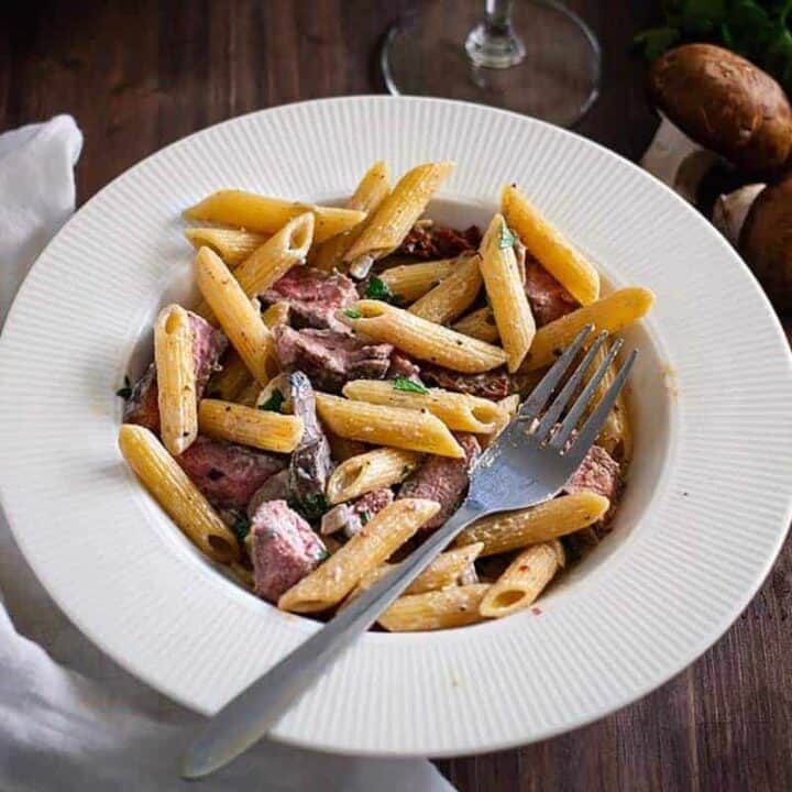 Creamy penne pasta with sliced prime rib on a white plate with a fork