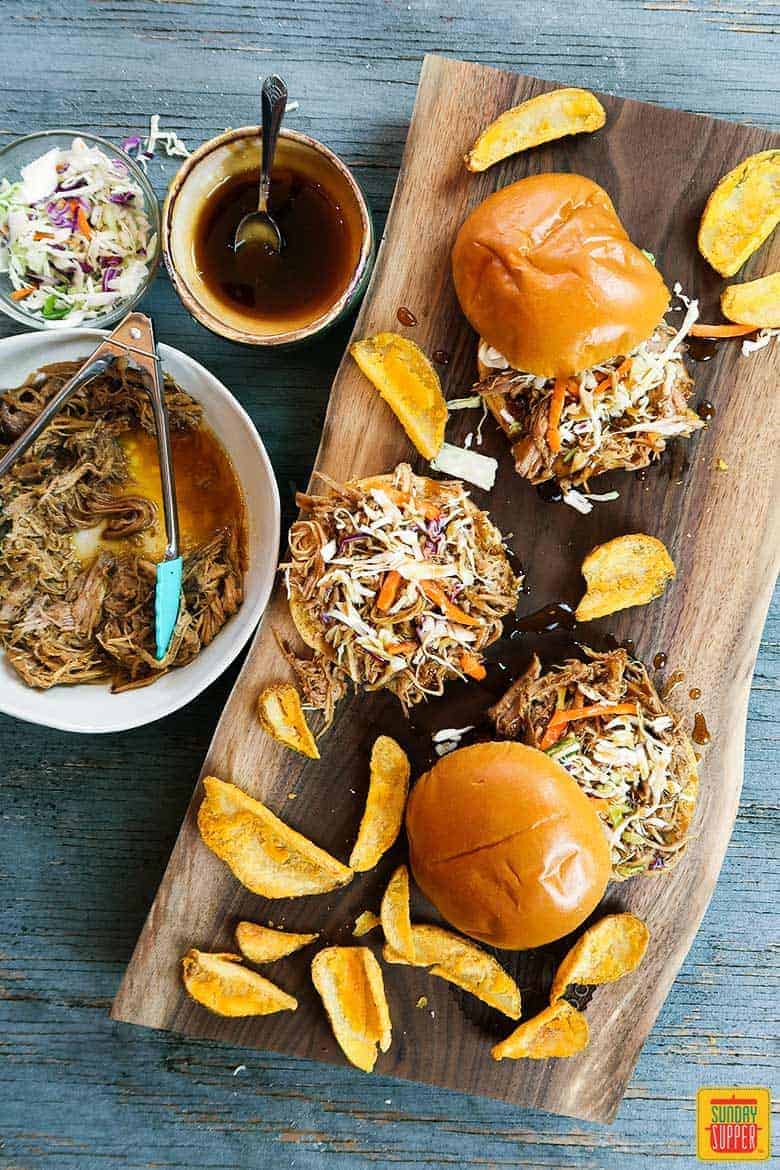 Three slow cooker pulled pork sandwiches on a cutting board with coleslaw and potato wedges