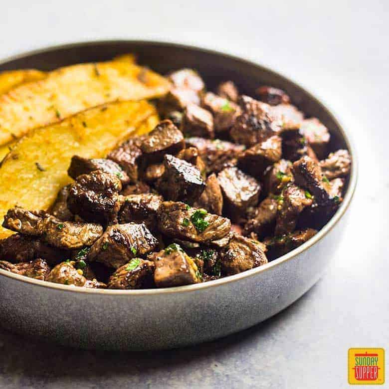 Garlic butter steak bites in a gray shallow bowl with potato wedges on the side for side dishes for steak