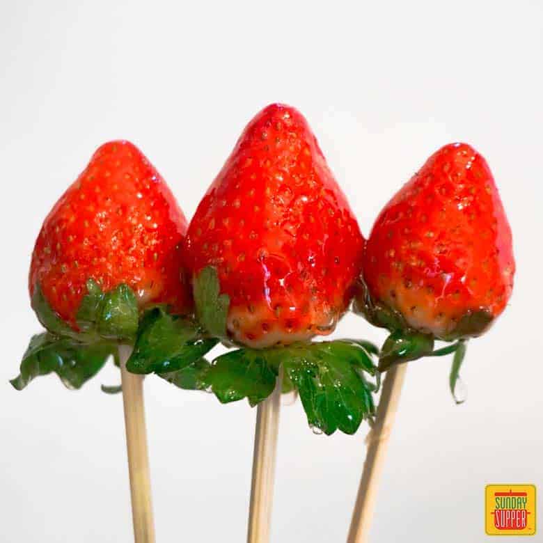 three candied strawberries on skewers over a white background