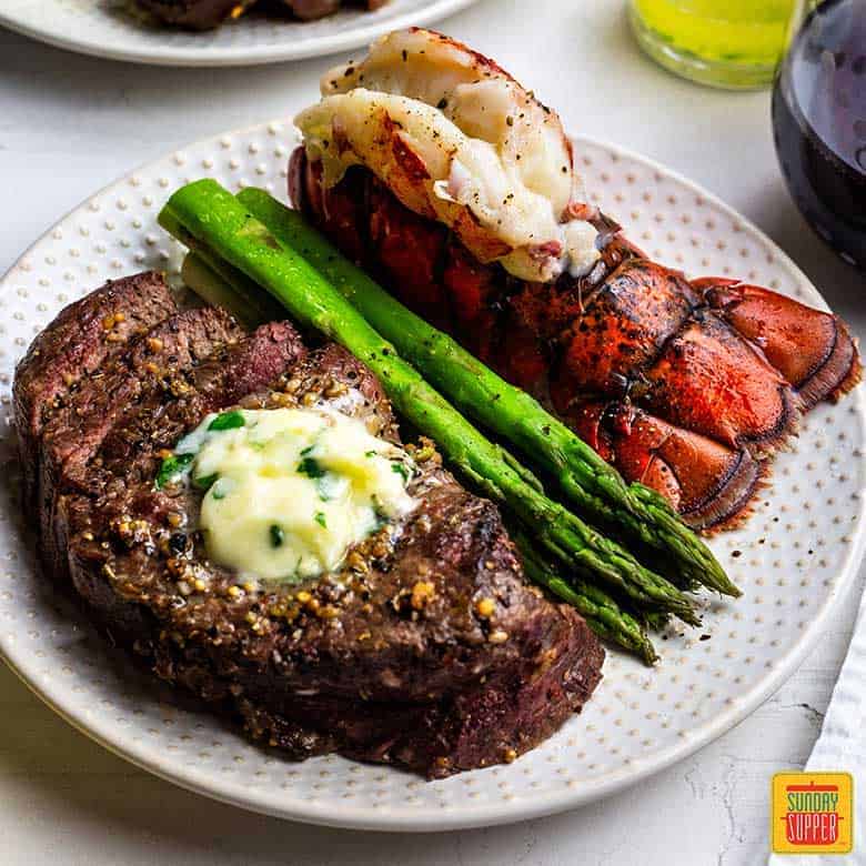 White plate with air fried steak and fried lobster tail with a side of steamed asparagus