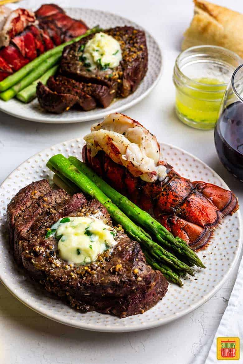 Two plates of surf and turf recipe with air fried steak, fried lobster tails, steamed asparagus, and garlic butter sauce for dipping