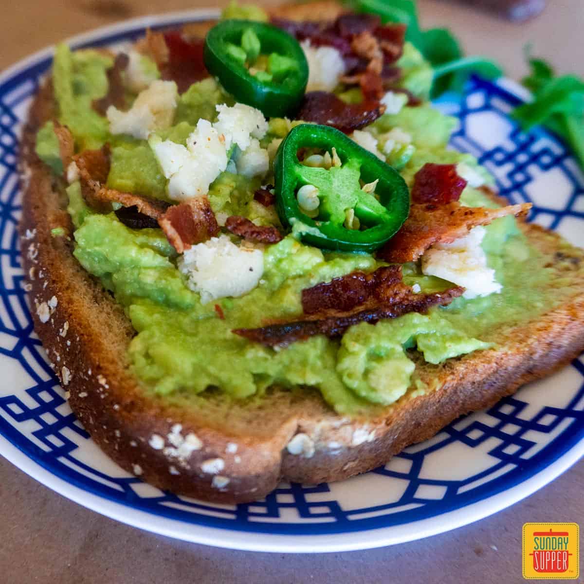 Two slices of avocado toast with bacon, goat cheese and jalapenos on a blue and white plate