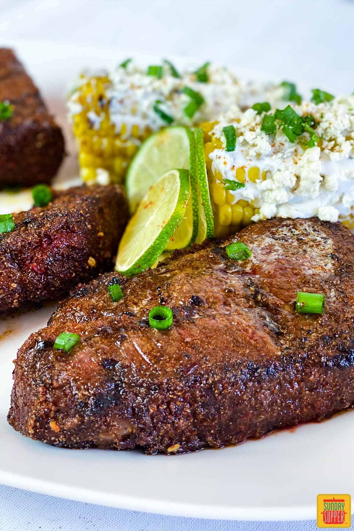 Close up of beef short ribs on a white plate with limes and elotes Mexicanos
