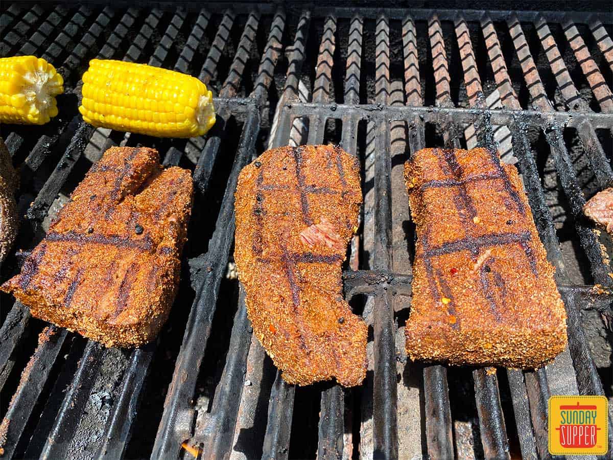 Close up of 3 beef short ribs on the grill with corn grilling alongside them