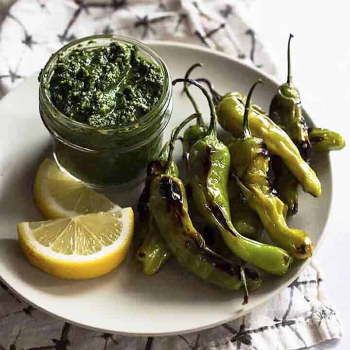 Blistered shishito peppers on a white plate with lemon wedges and citrus pesto dip in a glass jar