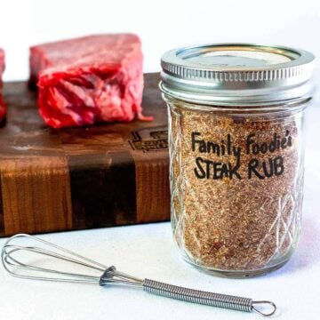 Close up of dry rub recipe in a glass jar in front of a beef short rib on a cutting board with a tiny whisk