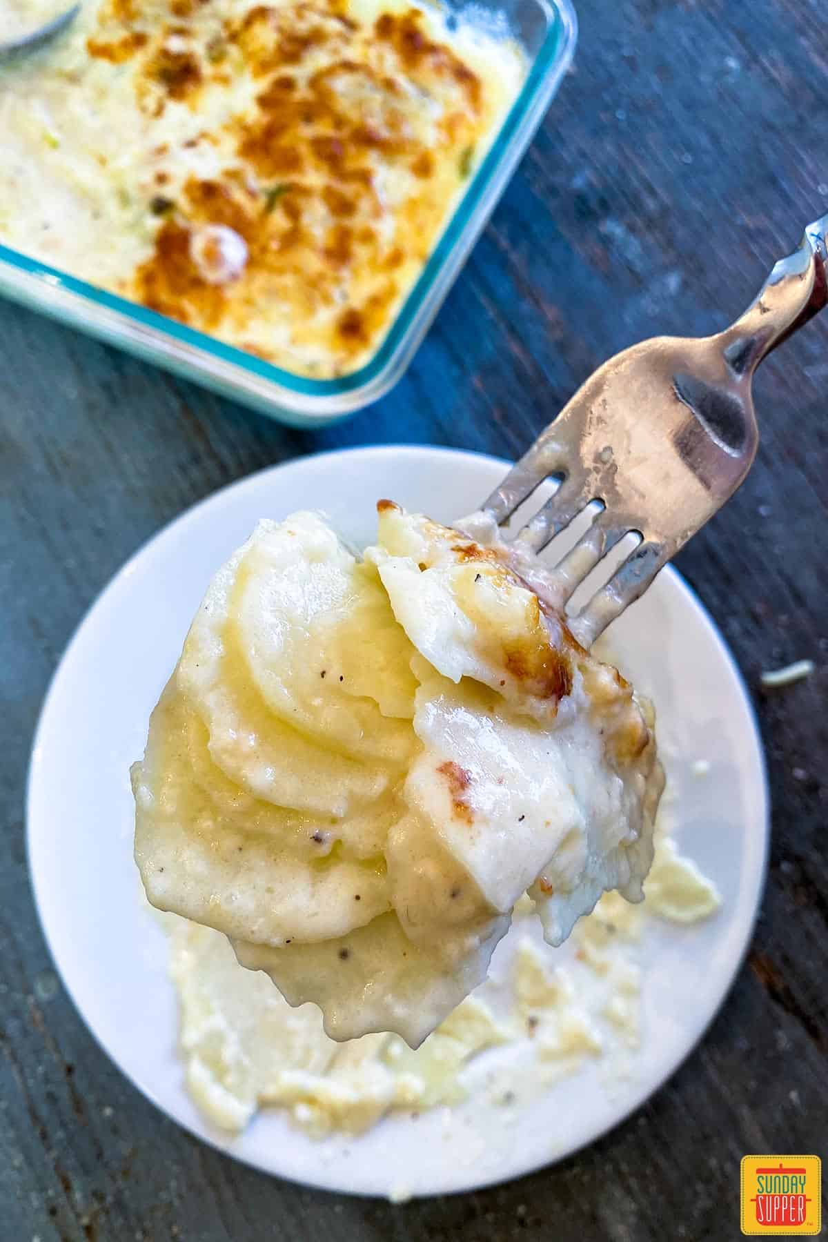 Holding a forkful of Instant Pot Scalloped Potatoes over a white plate near the casserole dish of potatoes