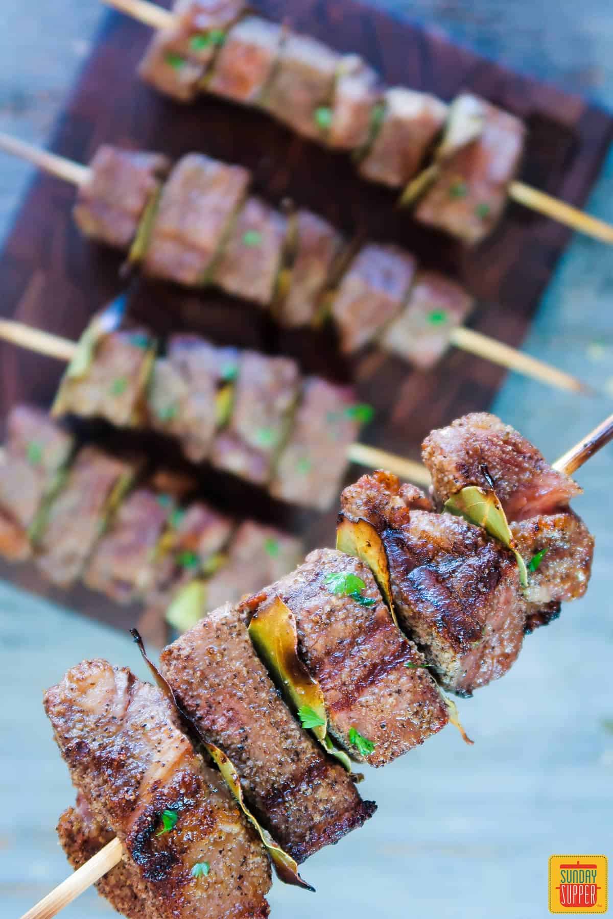 Holding up a Portuguese beef skewer after cooking it over a board of the other cooked skewers
