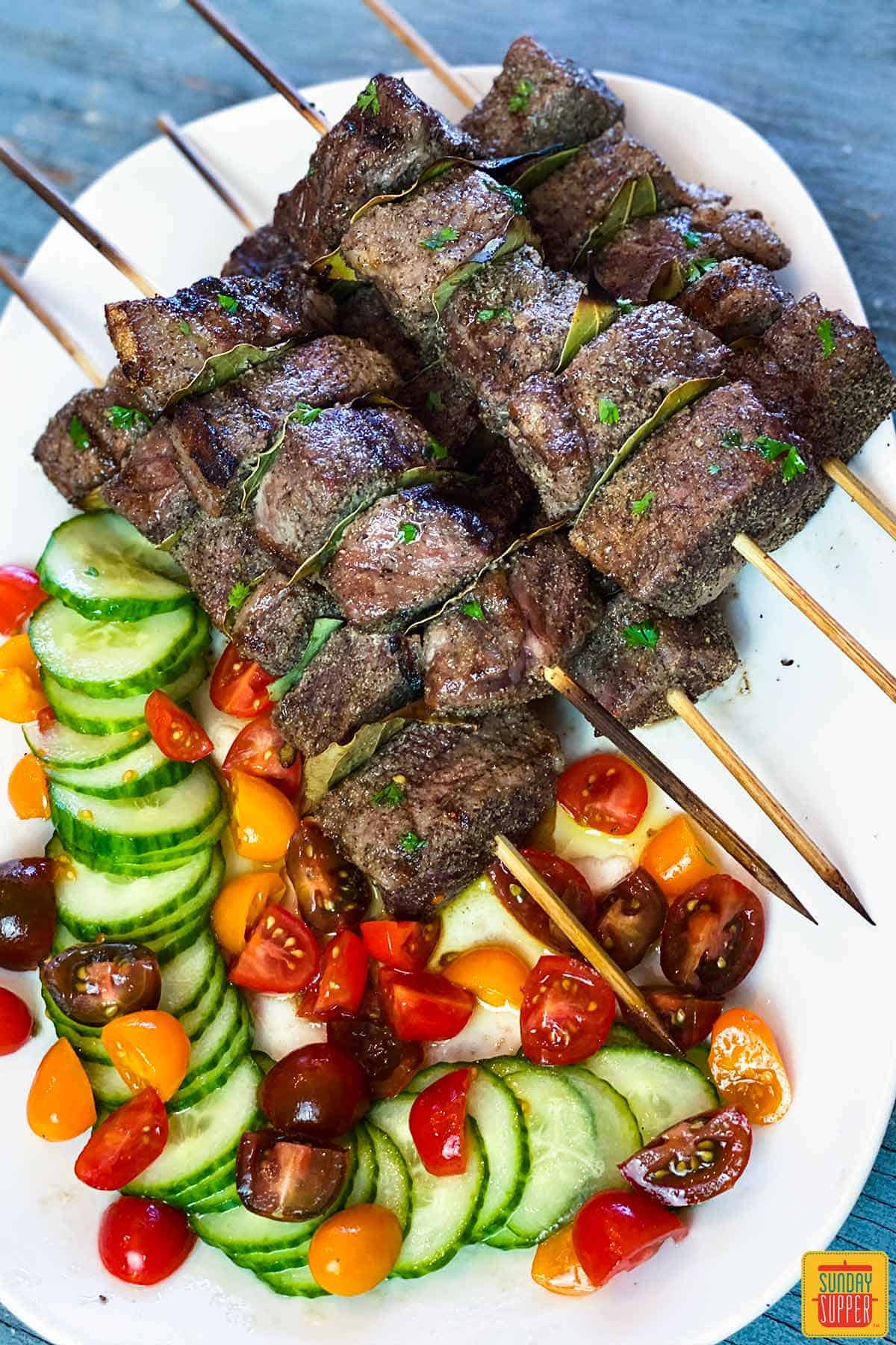 Overhead shot of Portuguese beef skewers on a white platter with tomato cucumber salad on the side