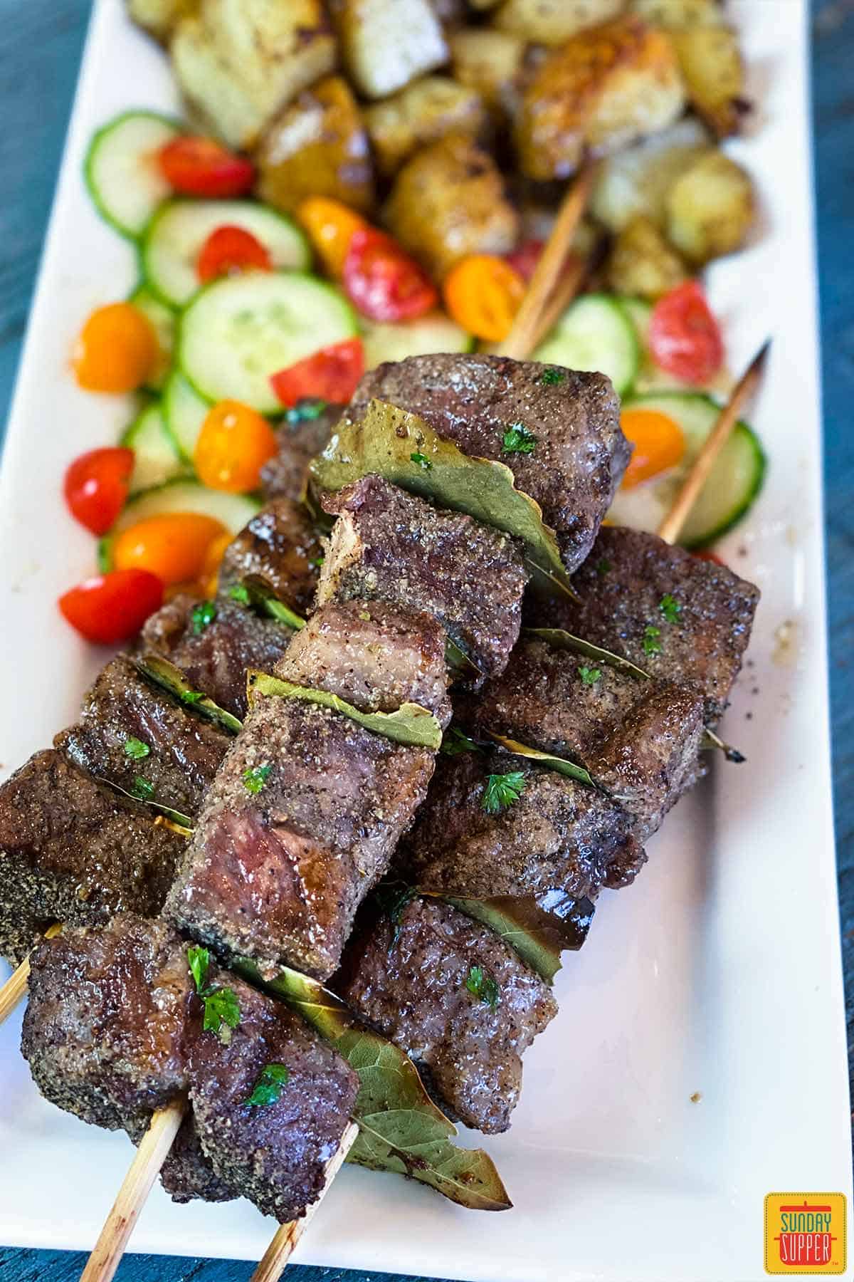 Close up of Espetadas Madeira on a plate with tomato cucumber salad and grilled potatoes