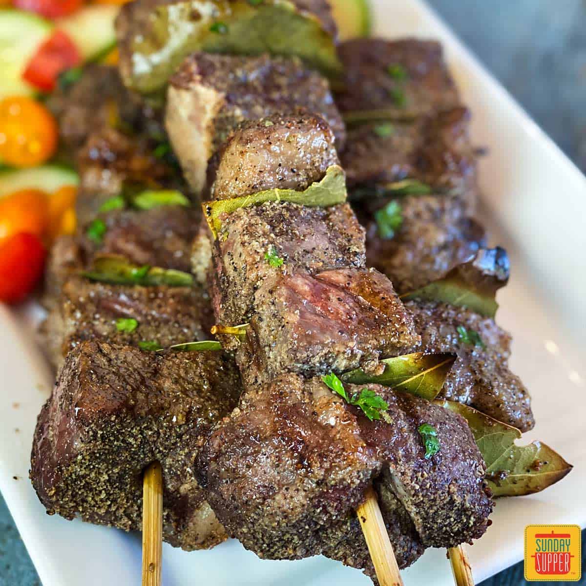 Close up of Portuguese Beef Skewers (Espetadas Madeira) on a white platter with tomato cucumber salad behind the beef