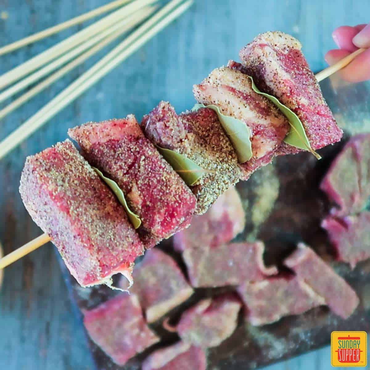 Holding a skewer layered with strip steak cubes and bay leaves in between each piece of steak