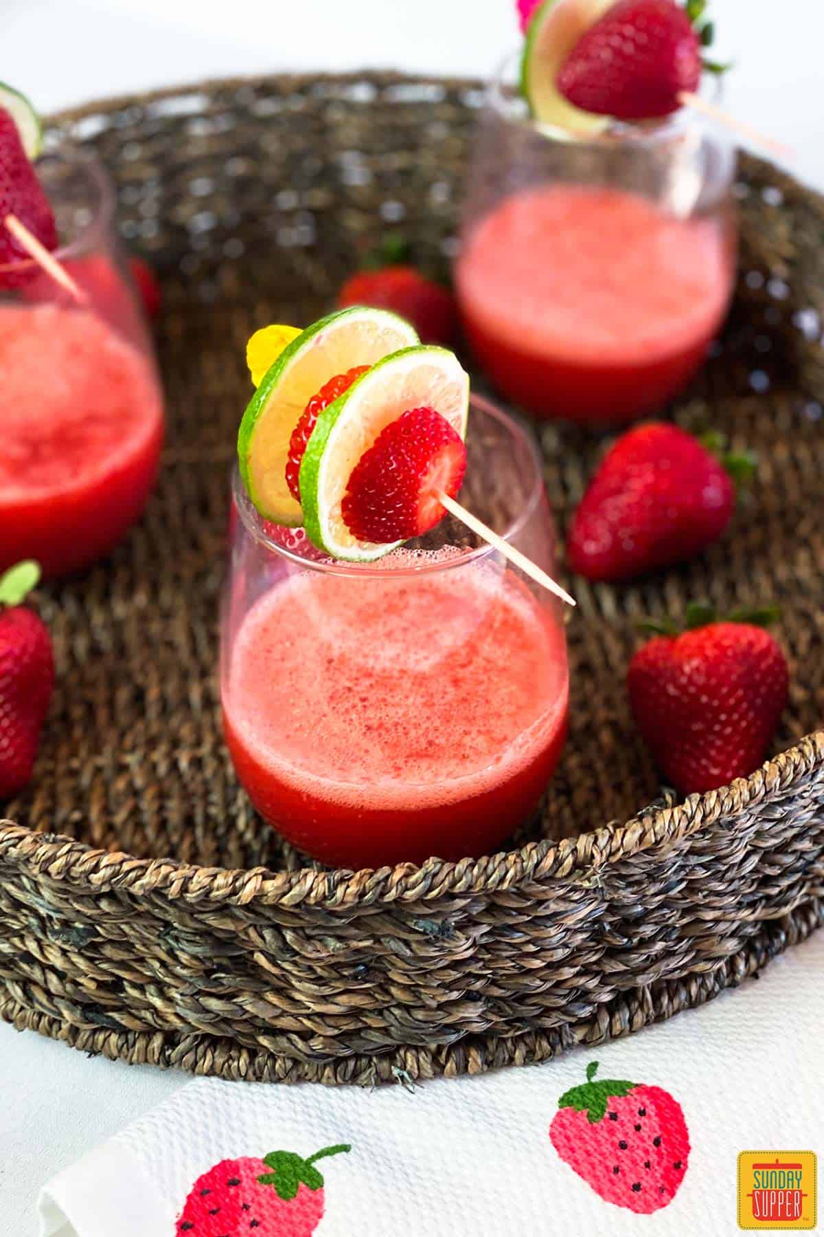 Three glasses of strawberry daiquiri in a flat-bottom round basket with fresh strawberries