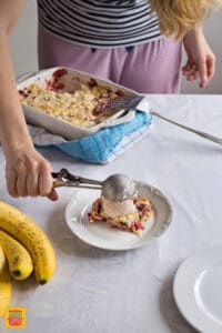A woman serving ice cream in a scoop over a slice of strawberry dump cake with bananas on a white decorative plate, with the whole dump cake in a baking dish in the background